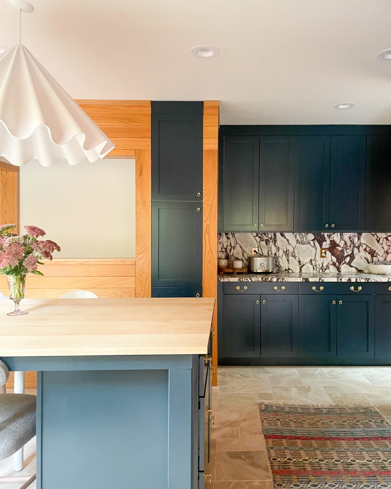 Kitchen dining area with navy blue cabinets, wood paneling, marble floors and countertops, and a vintage rug.