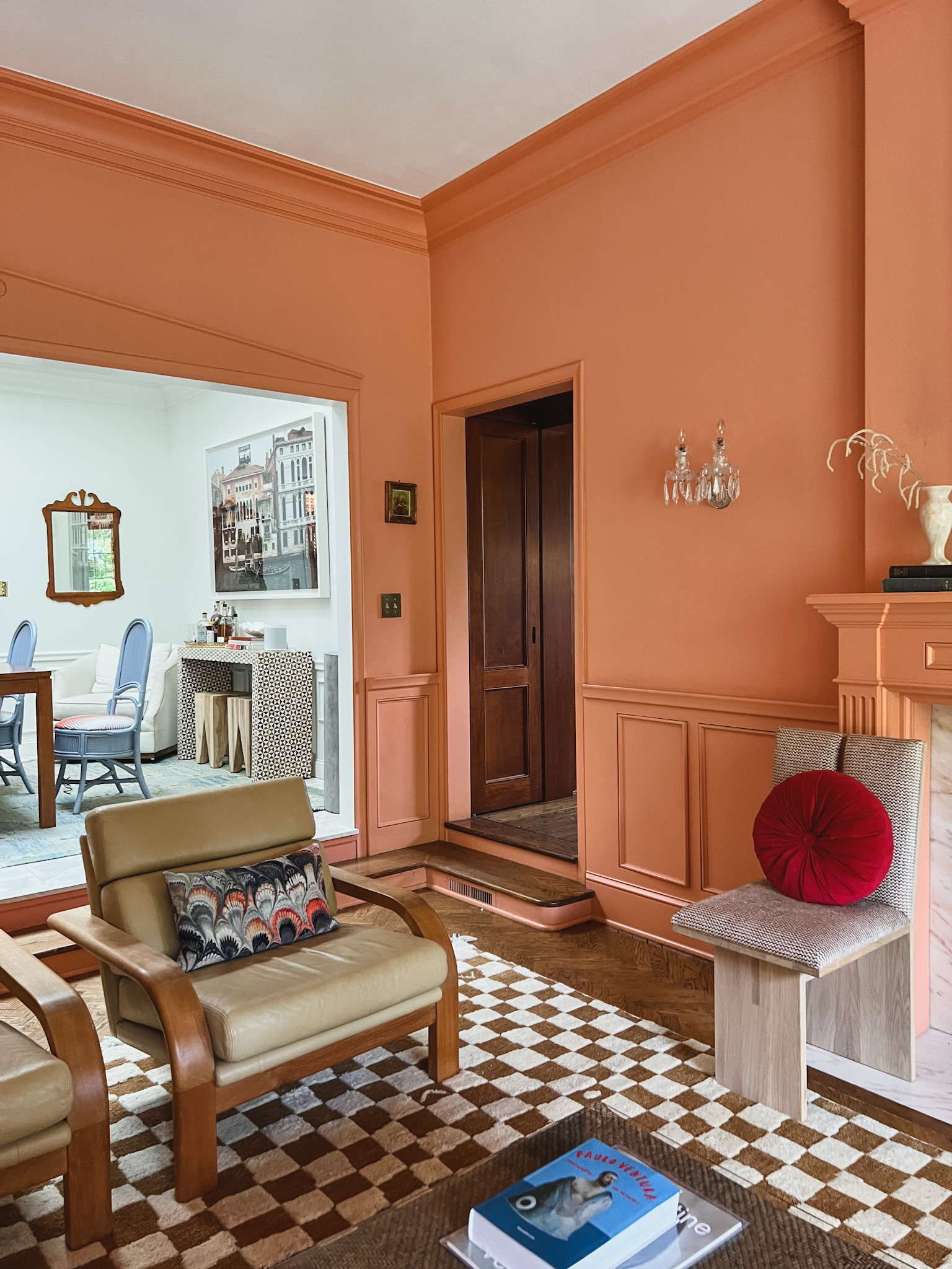 A brown and cream checkerboard rug grounds a living room with peach-colored walls, vintage chairs, and marbled accent pillows from Beata Hueman
