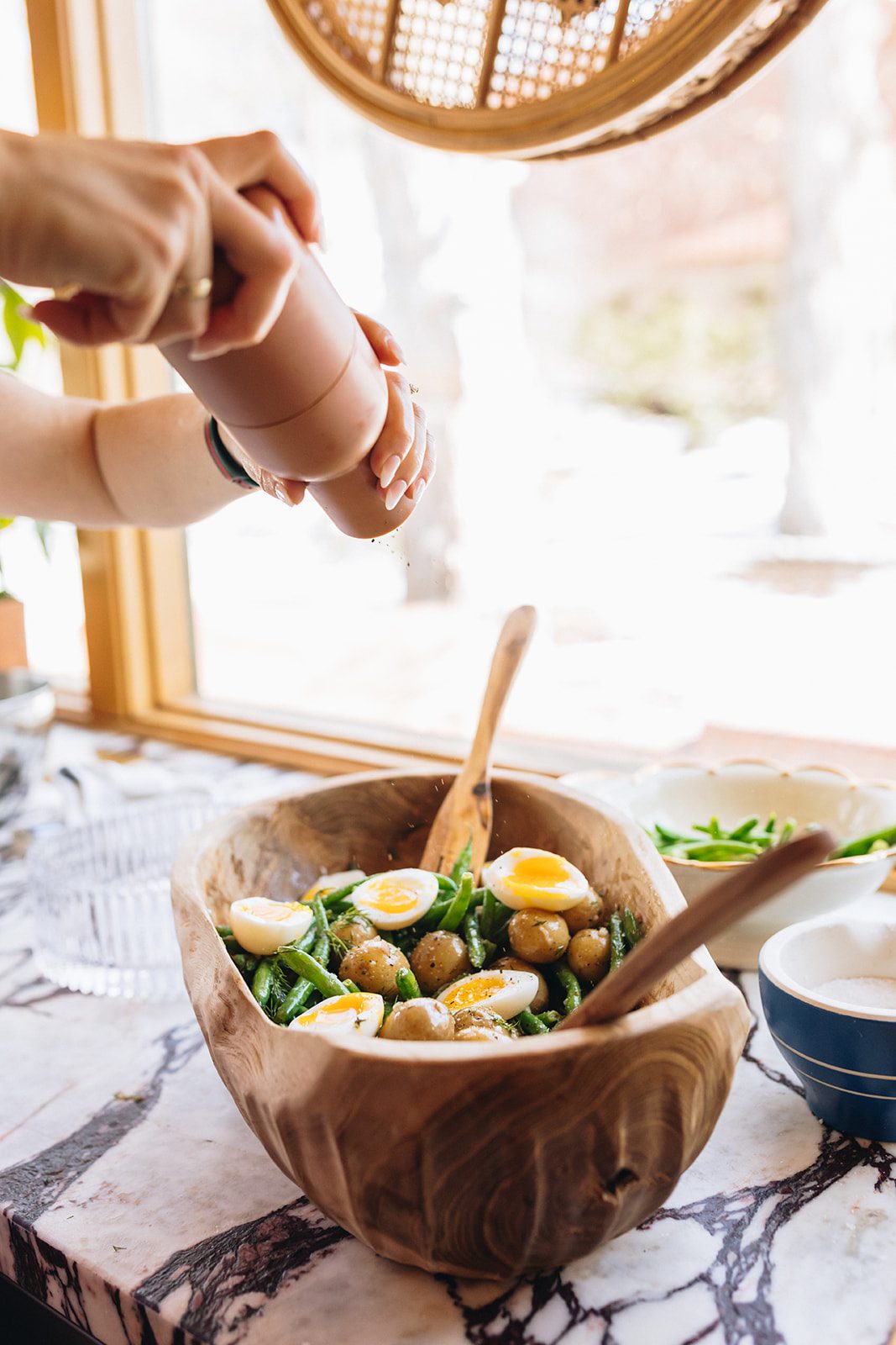 Hands cracking fresh pepper on top of a French green bean potato salad with medium-boiled eggs.