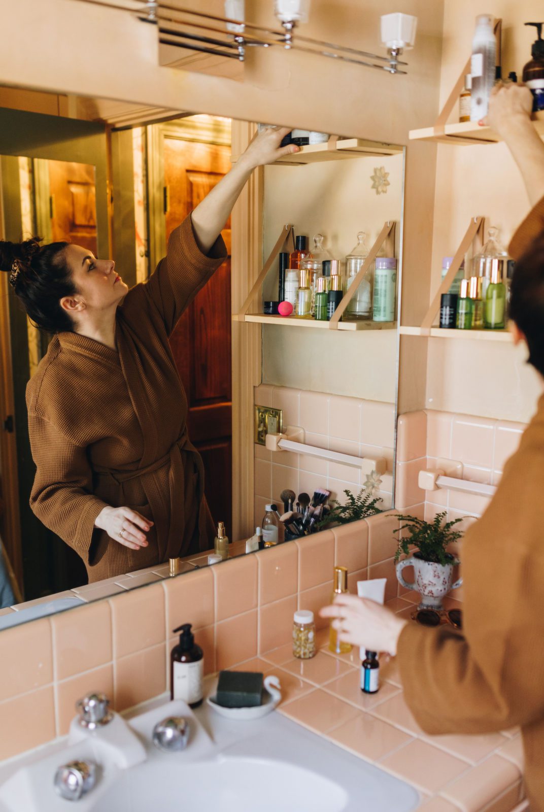 Woman reaching for skin-care product on top shelf of pink bathroom wearing a waffle robe. 