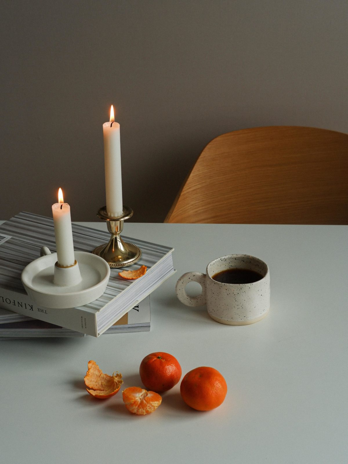 Taper candles lit on a white tabletop, with a cup of coffee and mandarin oranges.