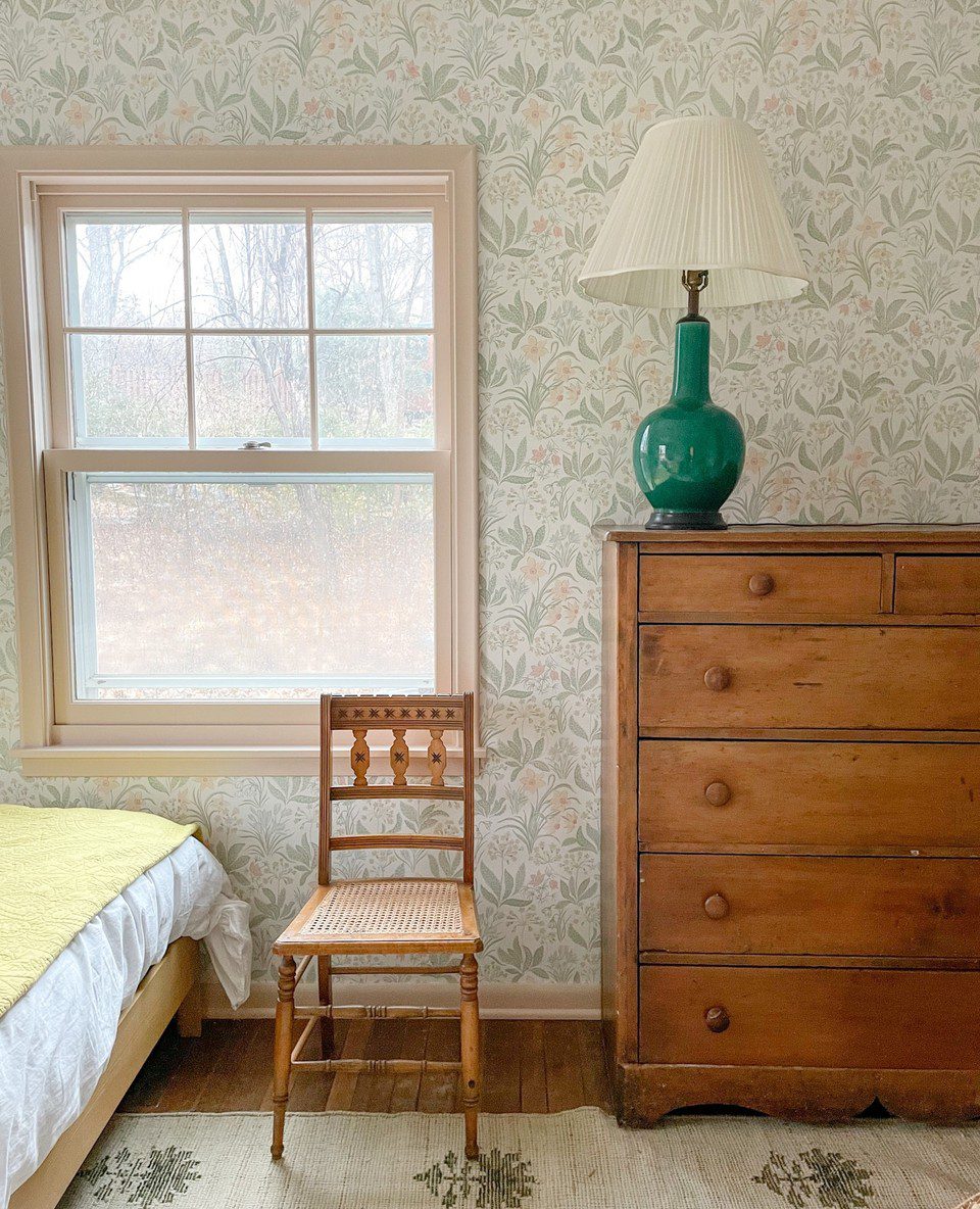 Wallpapered guest room with an antique dresser, table lamp, wooden chair, bed, and jute rug