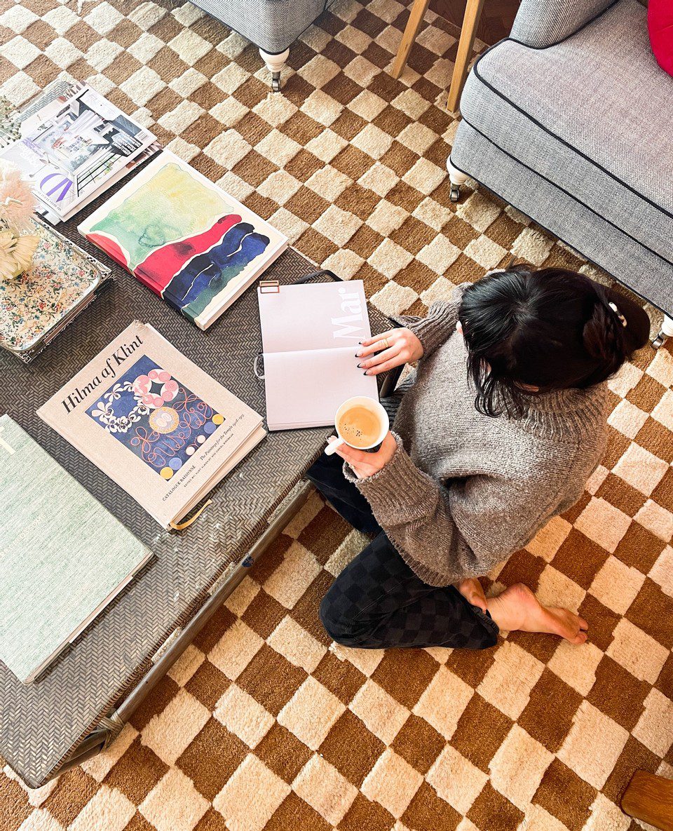 Woman sitting on a checkerboard rug paging through a planner with a cup of coffee in hand.