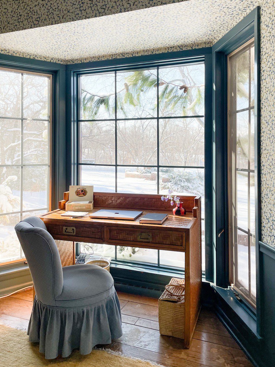 Antique wooden desk and blue ruffled chair in front of bay window in blue office.