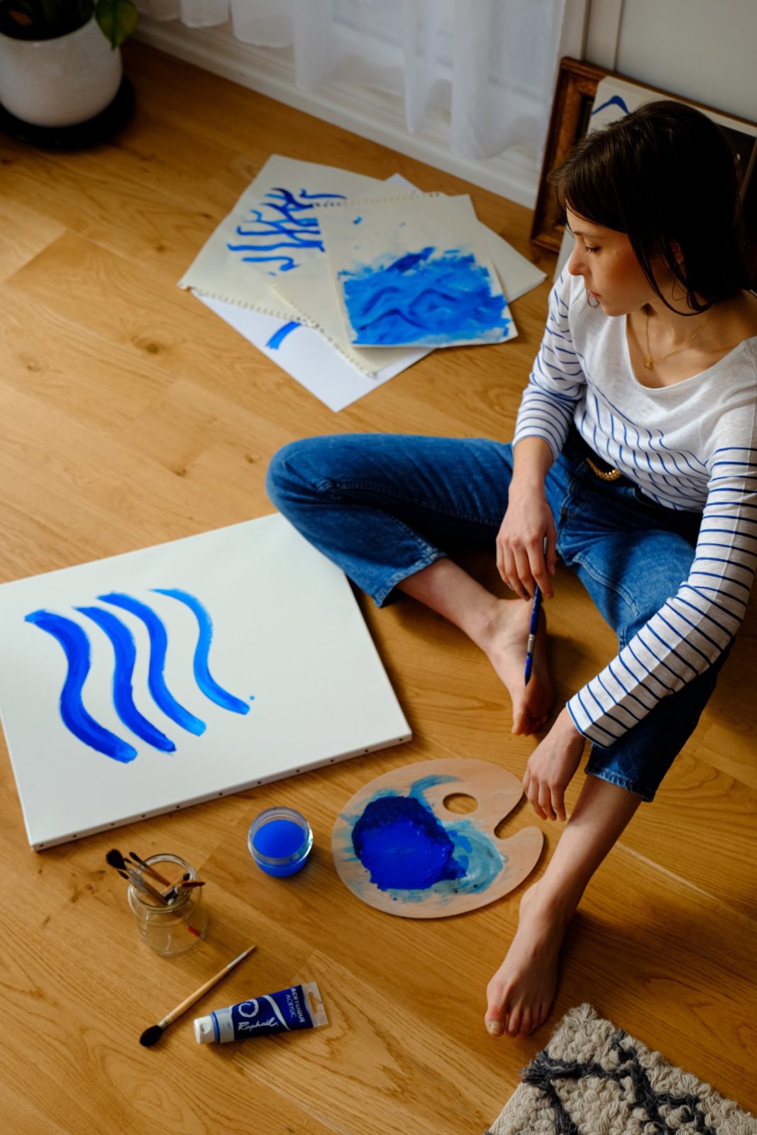 Woman sitting on a wood floor creating artwork with blue paint.