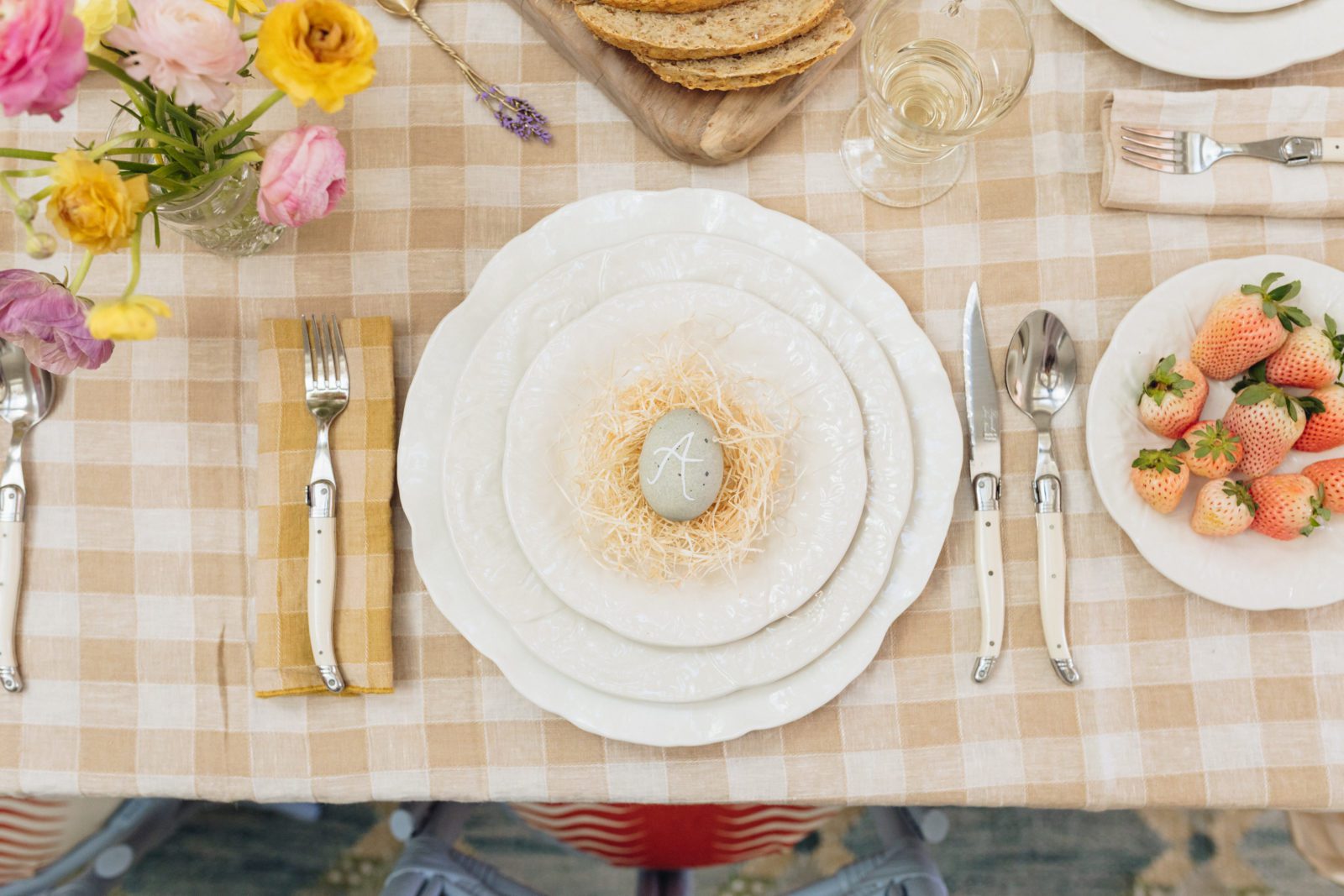 Top down view of a spring tabletop setting: charger, plate, fork, knife, spoon. On the plate is a handmade nest with a blue egg serving as a table setting.