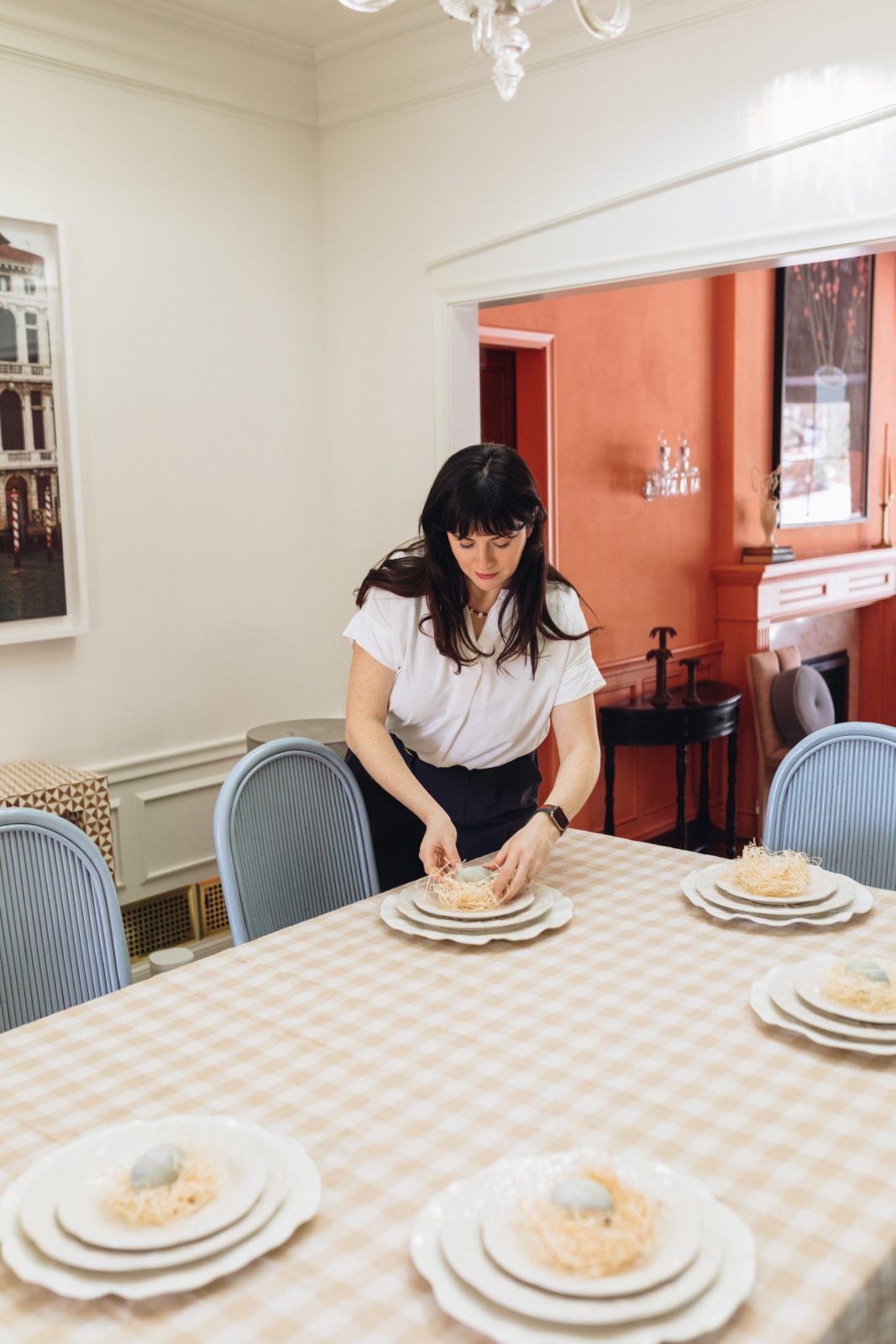 Woman setting a table in a bright and airy dining room. The woman is placing a handmade nest on top of a plate to serve as a placeholder.