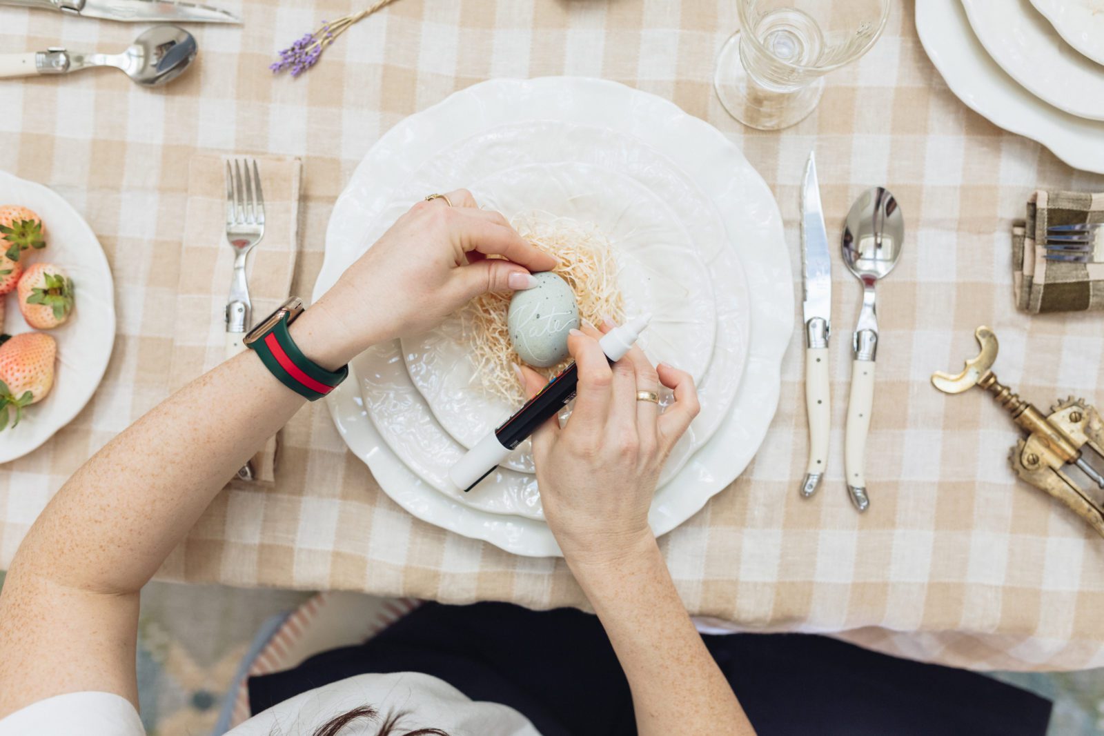 Top down view of a woman writing a name on a light blue egg. The egg is a seat placement for a semi-formal spring lunch.