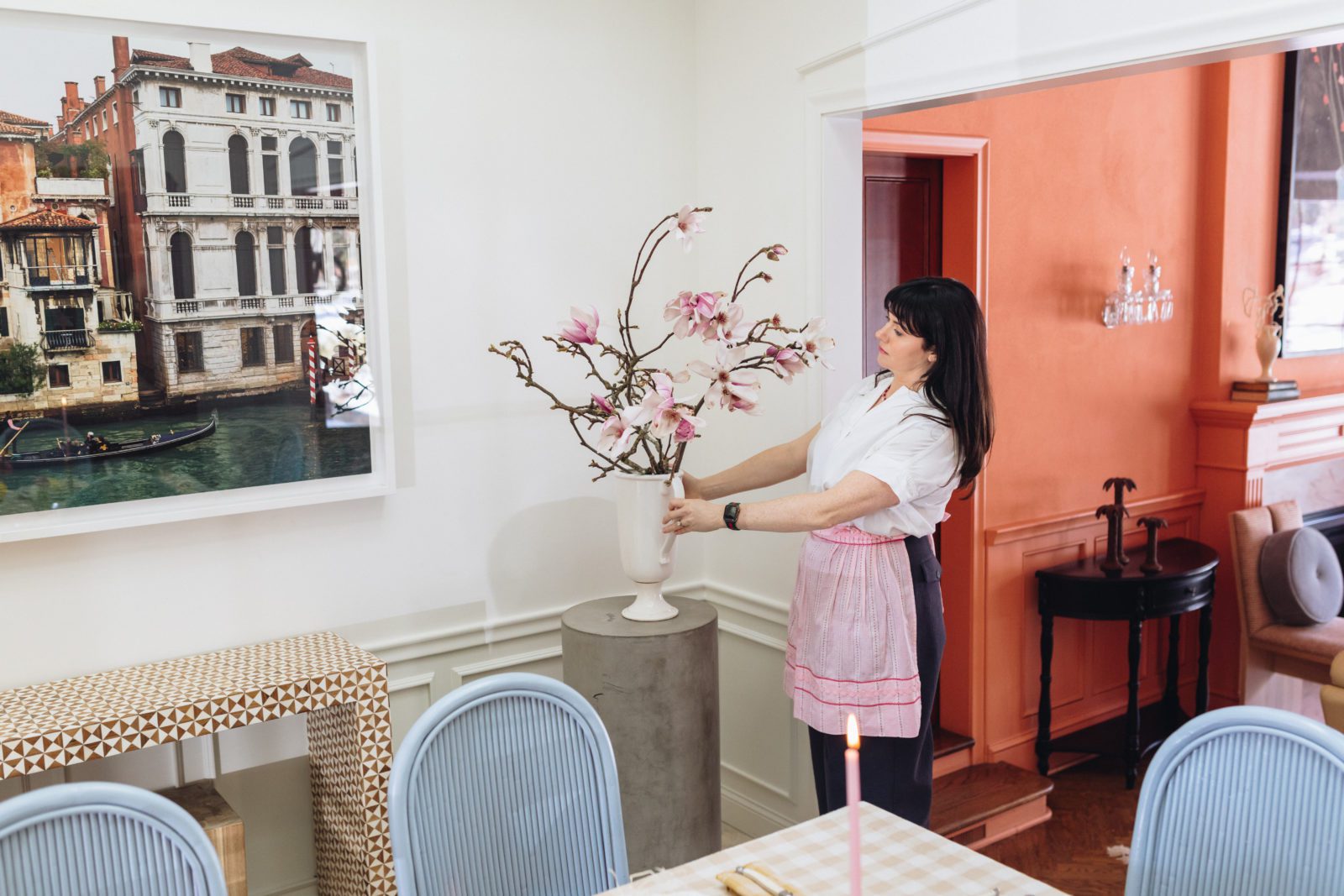 Woman arranging magnolia branches in a vase in a bright and airy dining room.