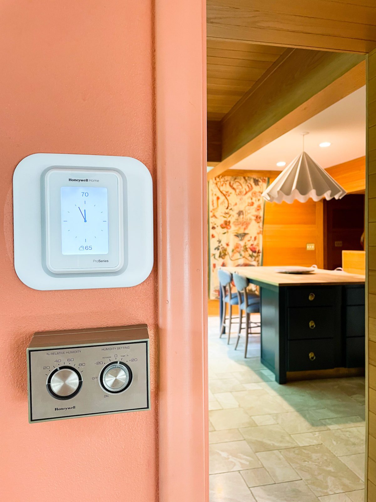 Close up image of a "smart" thermostat on the wall. In the background is a view of a kitchen.