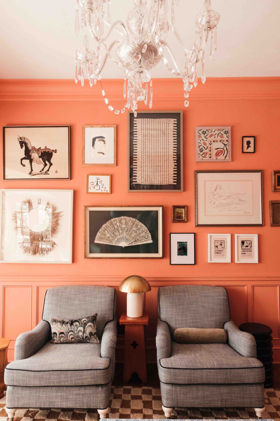 A living room features walls painted in a bold peach color, a large gallery wall, a set of armchairs with blue upholstery, contrast welting, and white turned legs, and a checkerboard rug