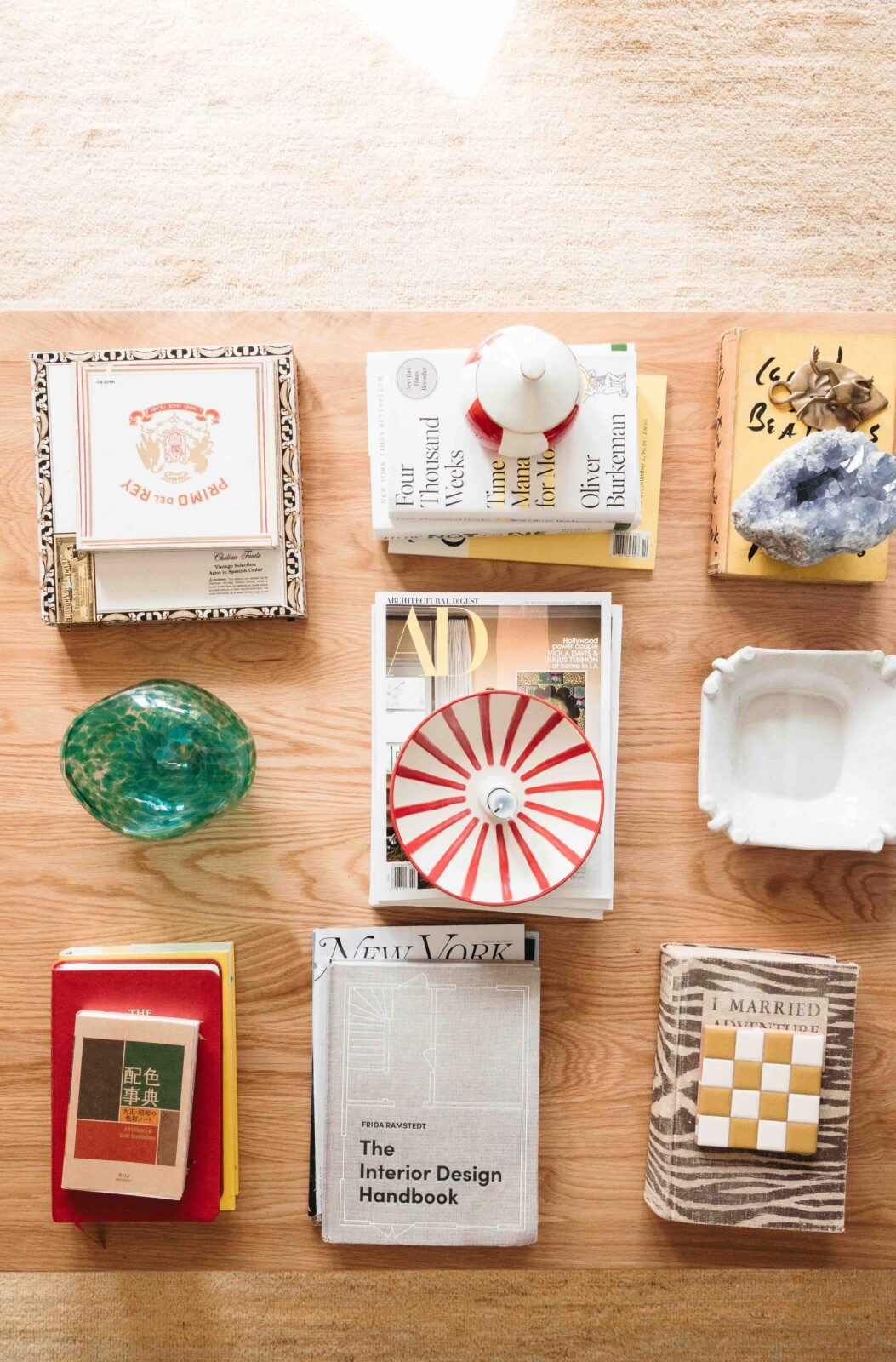 A square wood coffee table is topped with a variety of books, magazines, and decorative objects