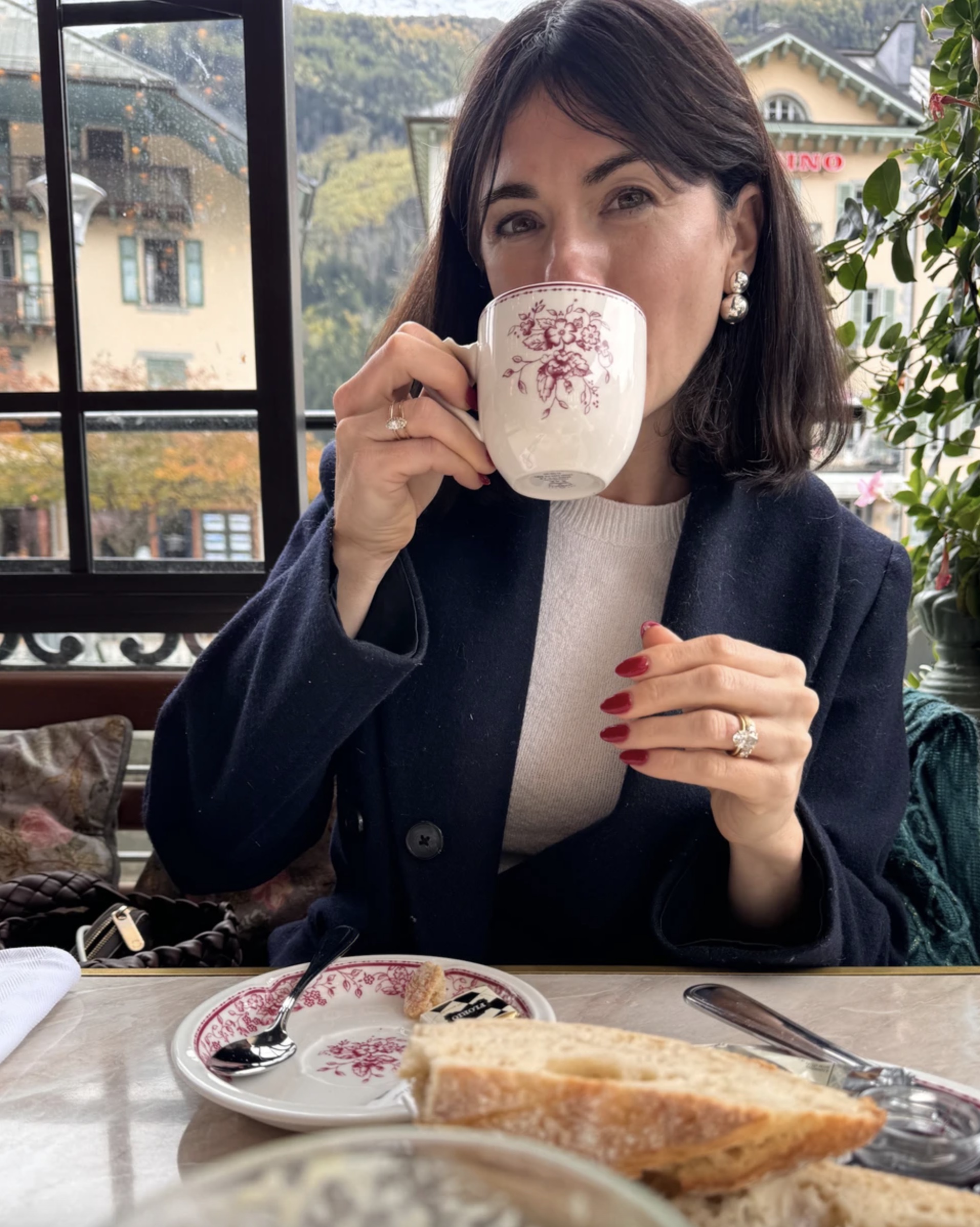 A woman wearing a navy wool coat and cream sweater, drinking a cup of coffee at lunch