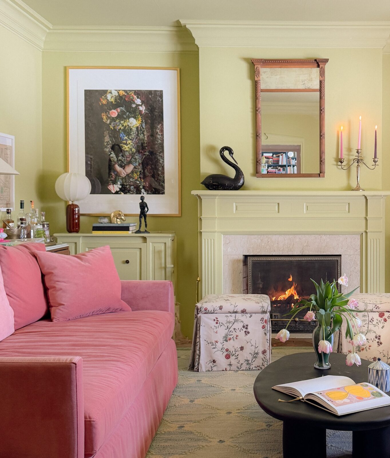 A green-painting family room with a pink velvet sofa, framed wall art, a black oval coffee table, floral upholstered ottomans, and a wood-burning fireplace