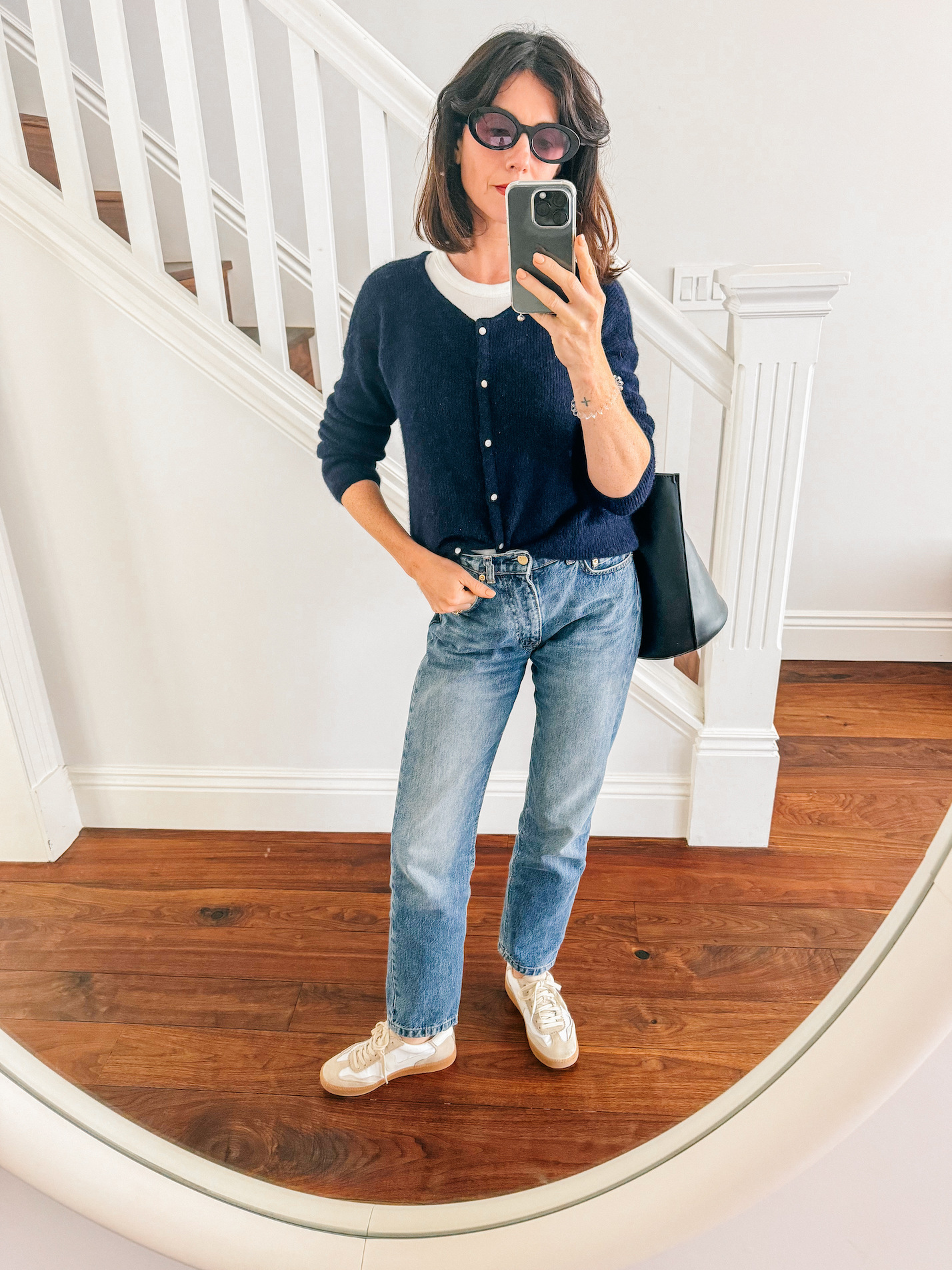 A woman stands in front of a staircase wearing blue denim jeans, ivory sneakers, a navy blue sweater, a white tank, and a black tote bag.