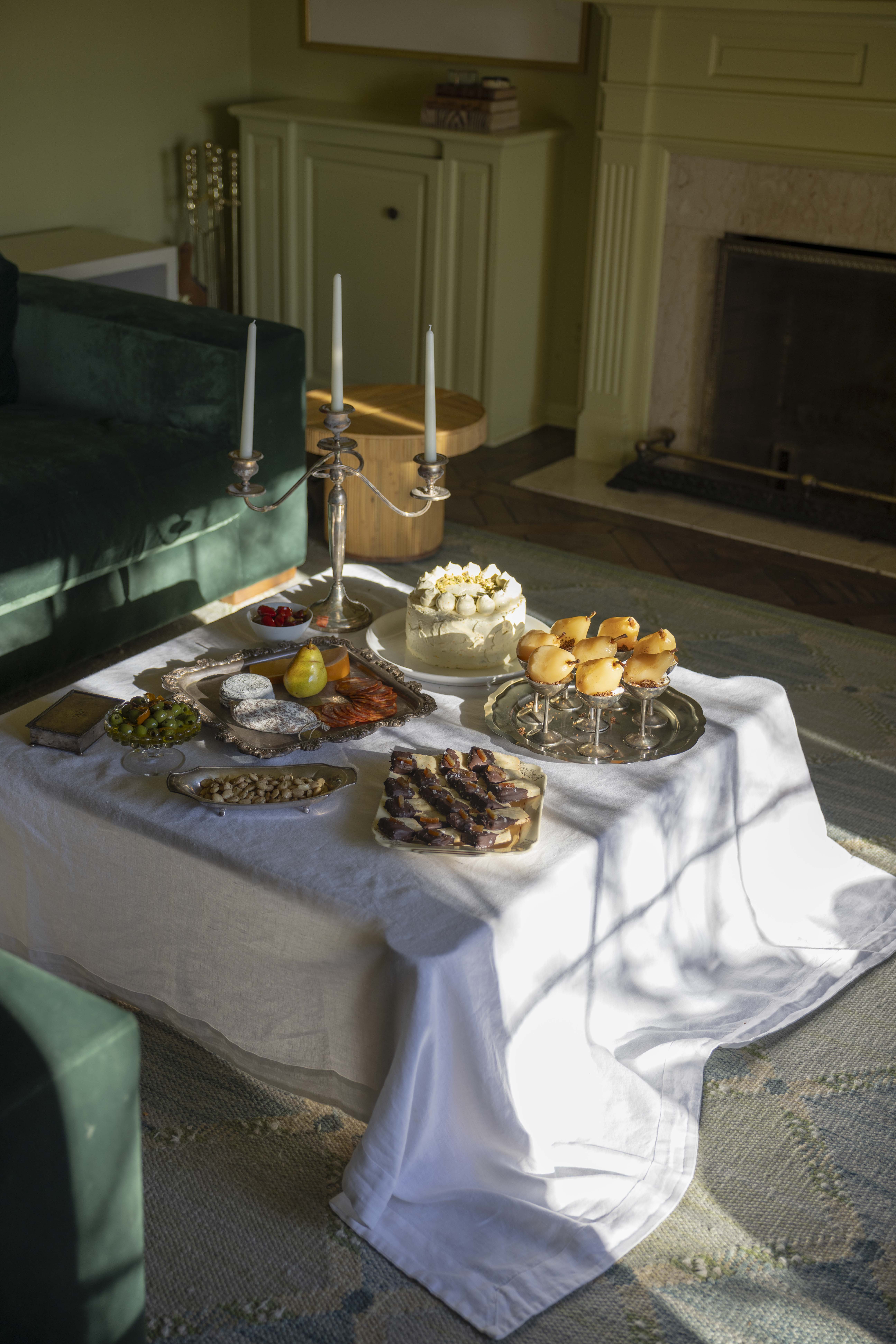 A coffee table is set for an afternoon dinner party. Serving trays hold shortbread cookies, ricotta olive oil cake, poached pears, and charcuterie