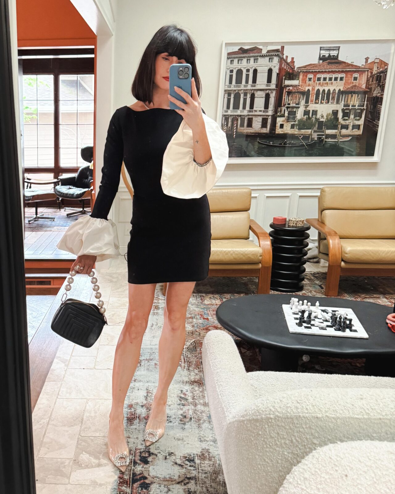 A woman stands in front of a full-length mirror, dressed for a wedding in a black mini dress with white bell sleeves, a black bag with pearl handle, and clear heels with pointed toes