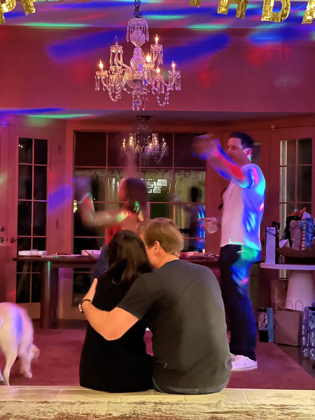 A couple sits on the steps in their home during a party, while disco lights cover the walls and people dance in the background