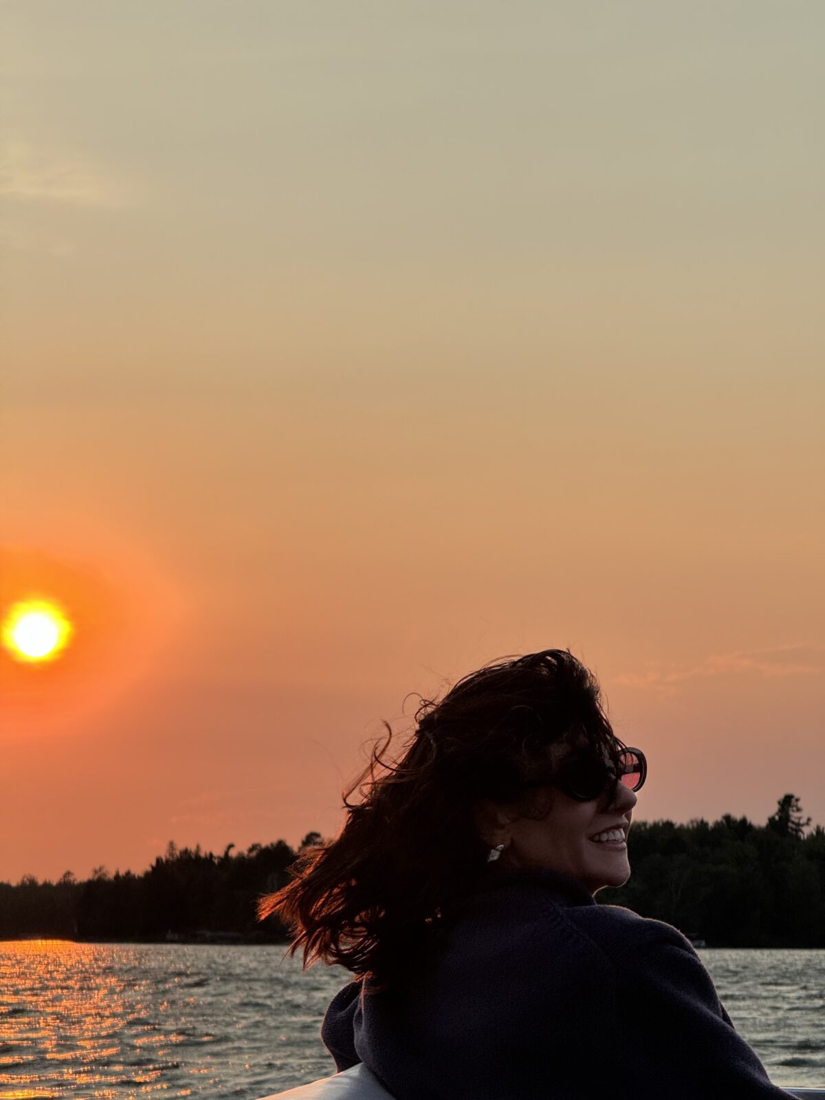 A woman sits on a boat in northern Minnesota, smiling as her hair blows in the wind and the sun sets behind her