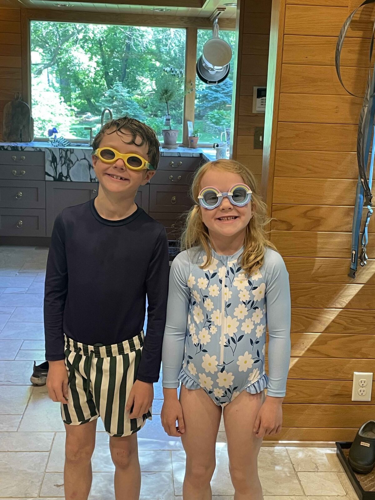 Two kids stand in their kitchen in the summertime, dressed for the pool wearing swimsuits and goggles