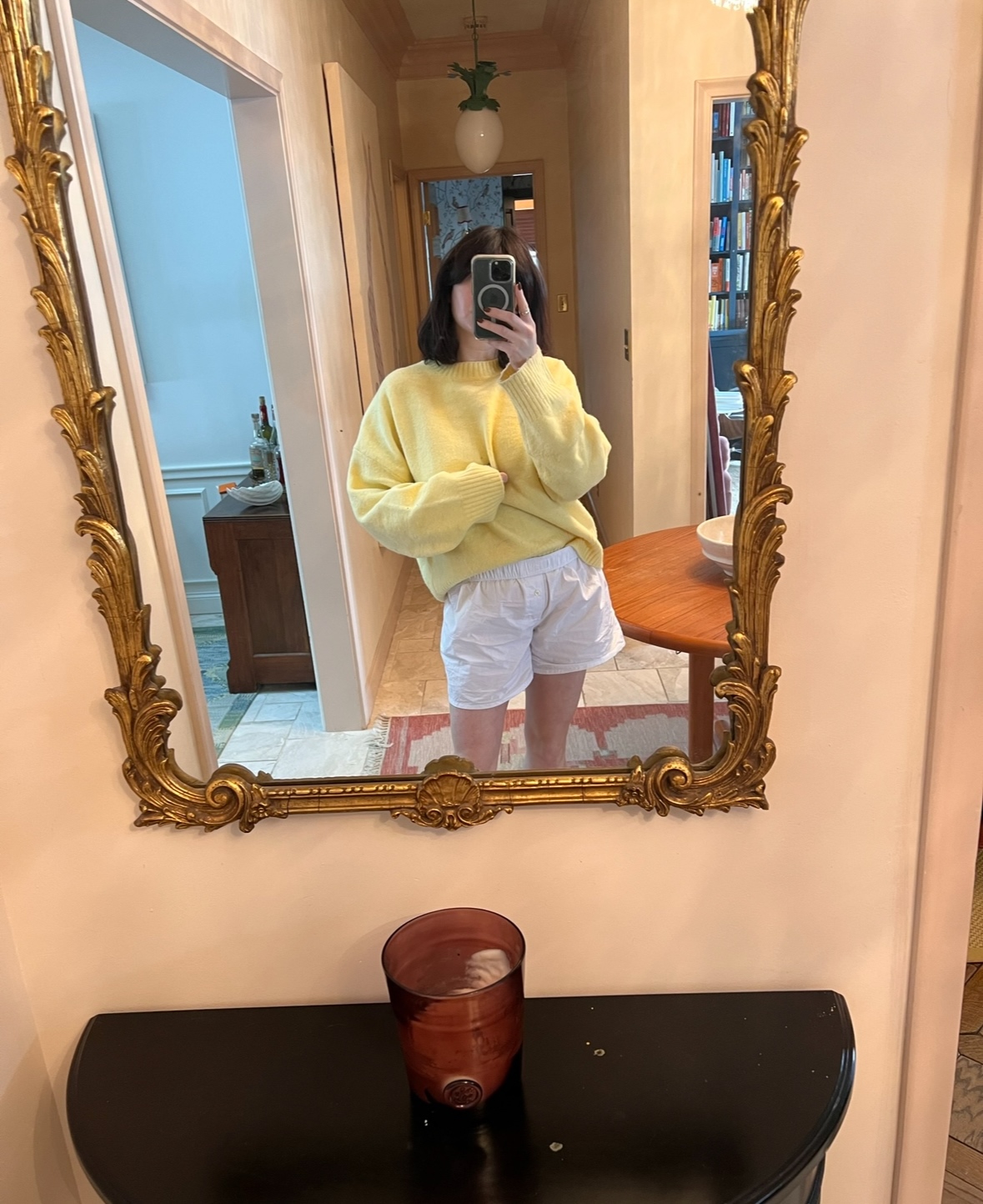 A woman stands in front of a vintage mirror in her entryway, wearing white linen shorts and a cozy, buttery yellow crewneck sweater