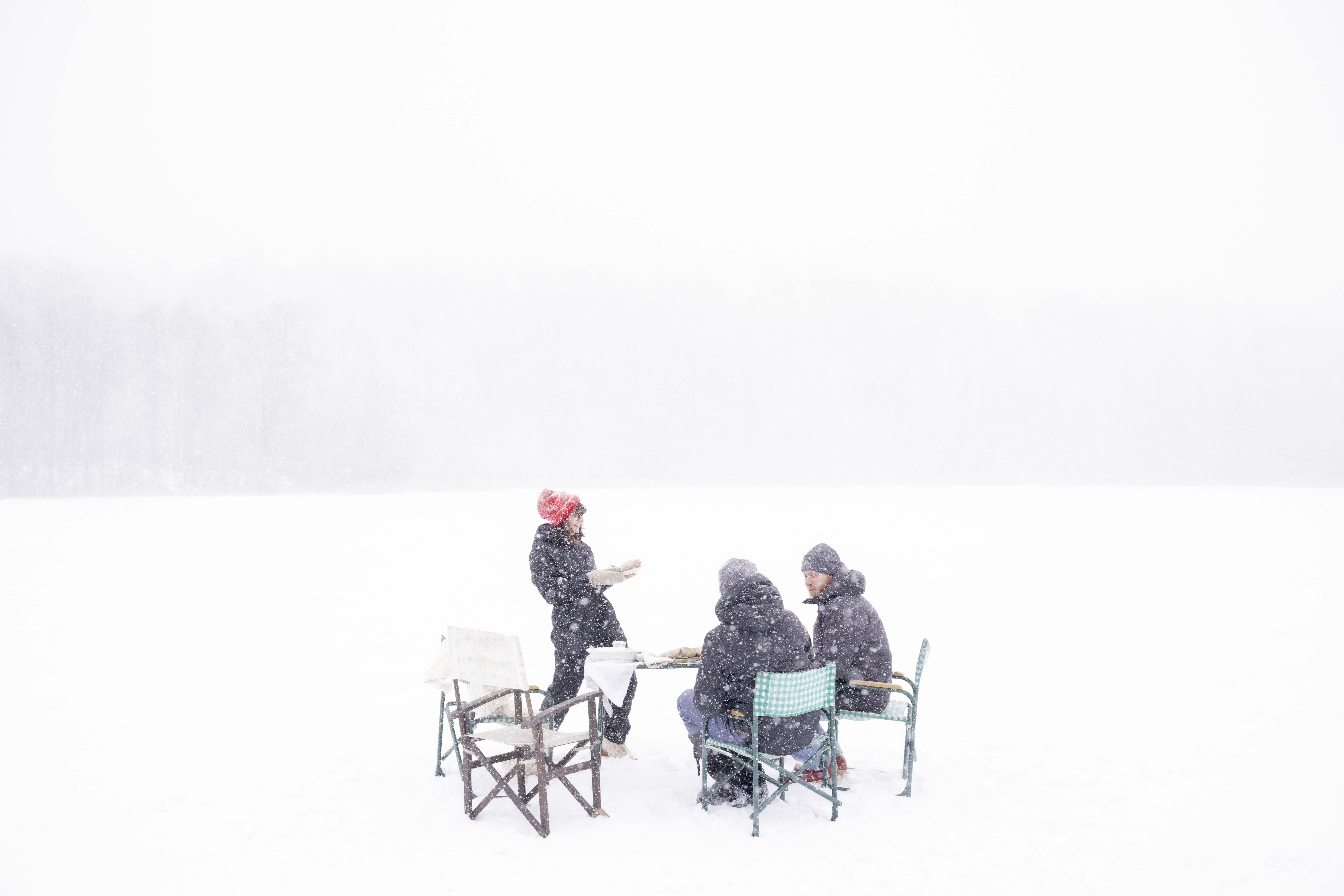 In the middle of an ice-covered lake in Minnesota on a snowy day, friends gather around a table for a homemade lunch