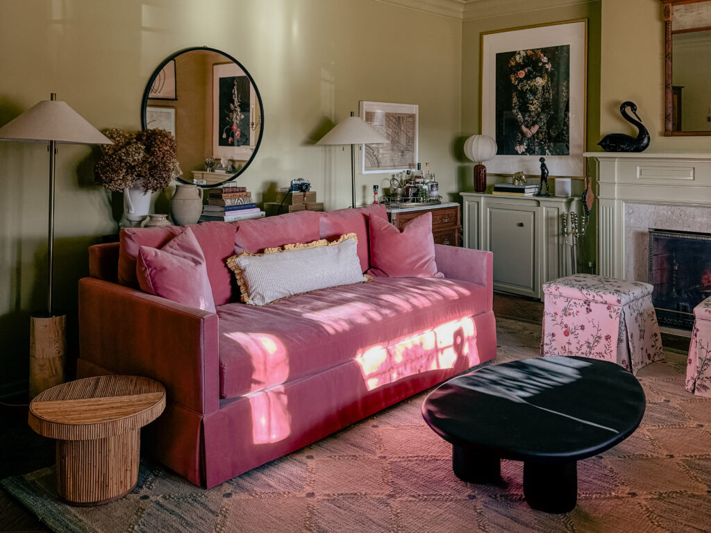 A green-painted living room features a pink velvet sofa and floral upholstered ottomans from The Inside, a low black coffee table, and a blue and green, diamond-patterned rug