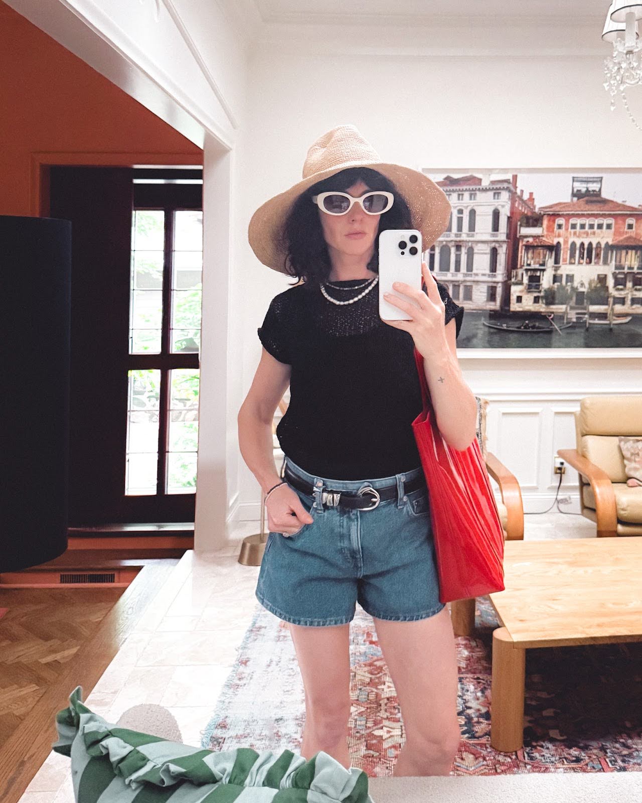 A woman wears denim shorts, and black and silver belt, a black sweater tee, a white sunglasses, a big sun hat, and a red patent leather bag