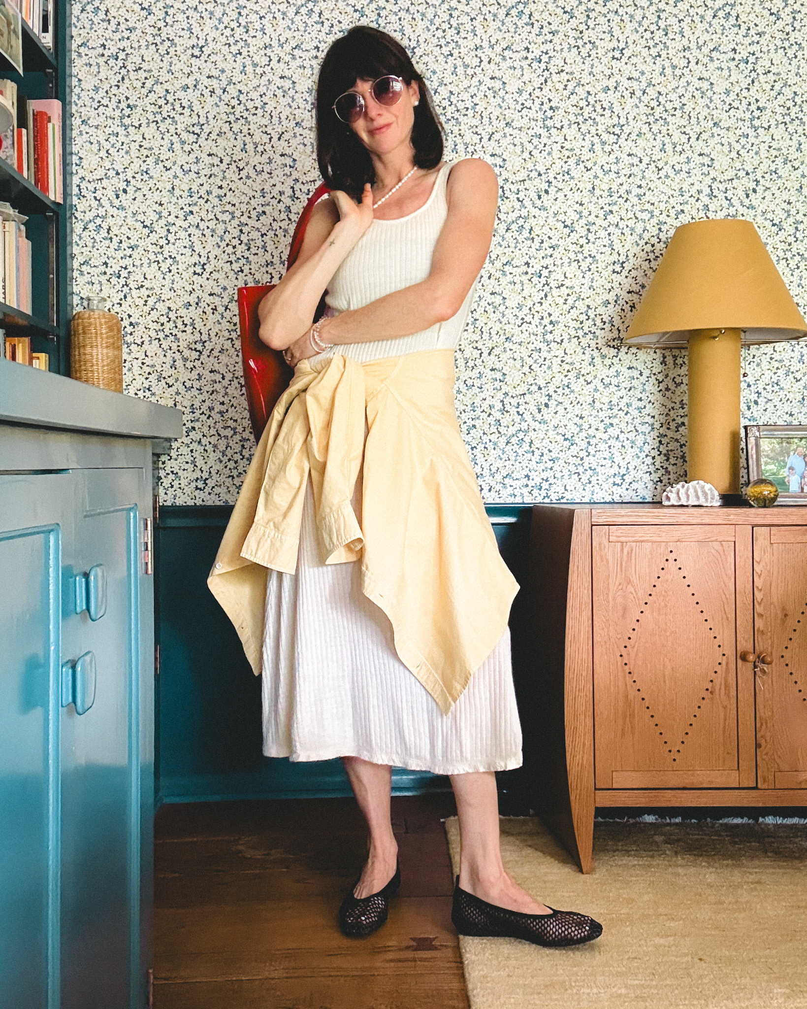 A woman wears a long white knit dress, a yellow button-down shirt tied around her waist, black ballet flats, sunglasses, and a red patent leather bag