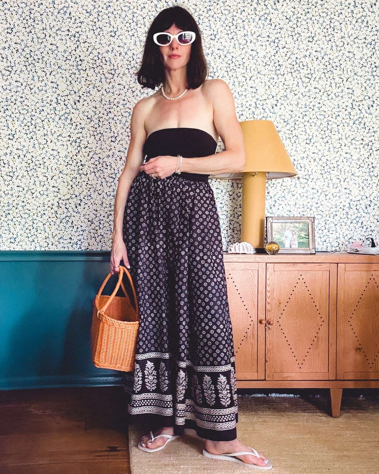 summer outfit ideas: A woman wears a strapless maxi dress, white flip flops, and sunglasses while holding a brown basket bag