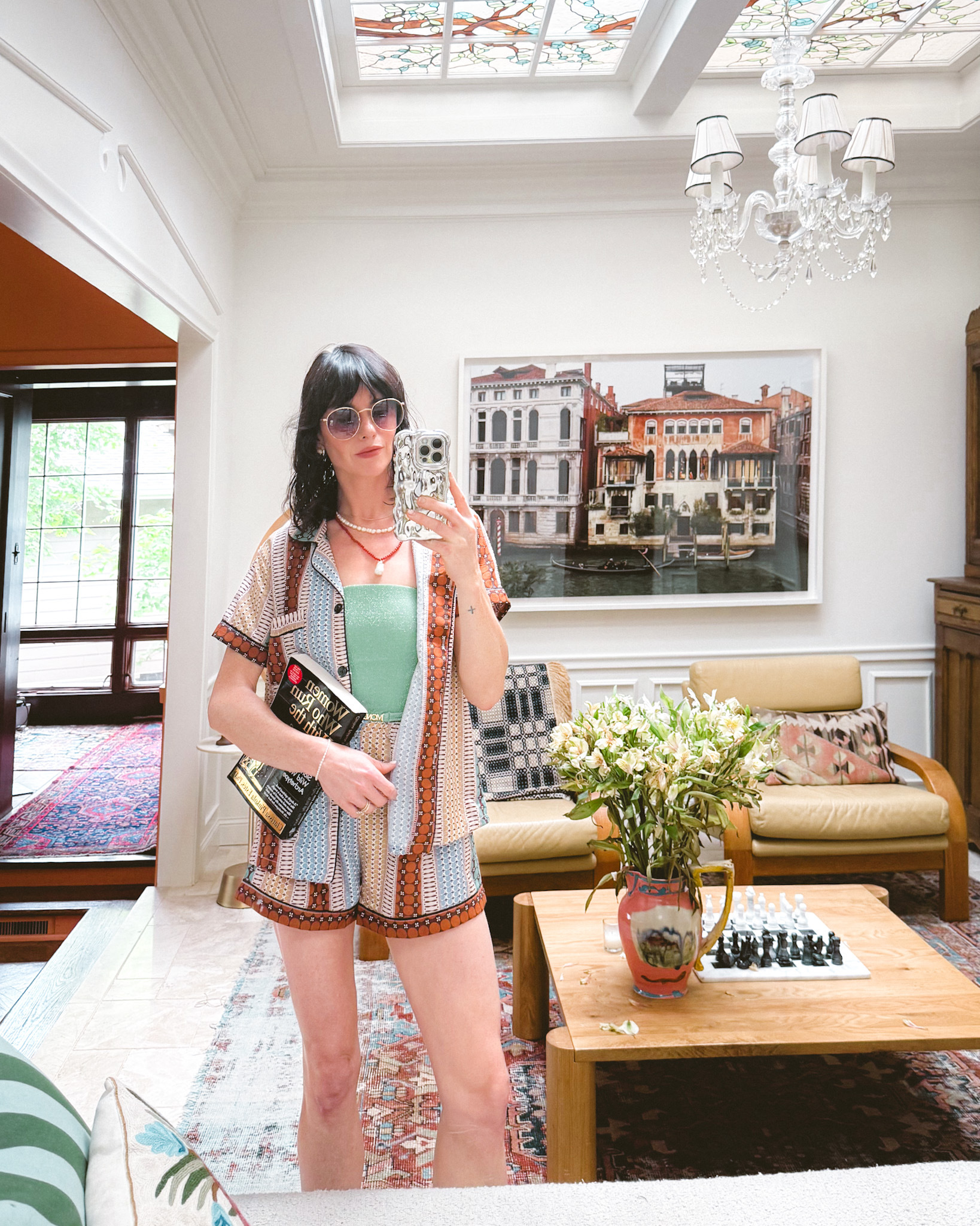 Summer outfit ideas: a woman wears a green swimsuit, a patterned shirt and shorts set, sunglasses, and layered necklaces