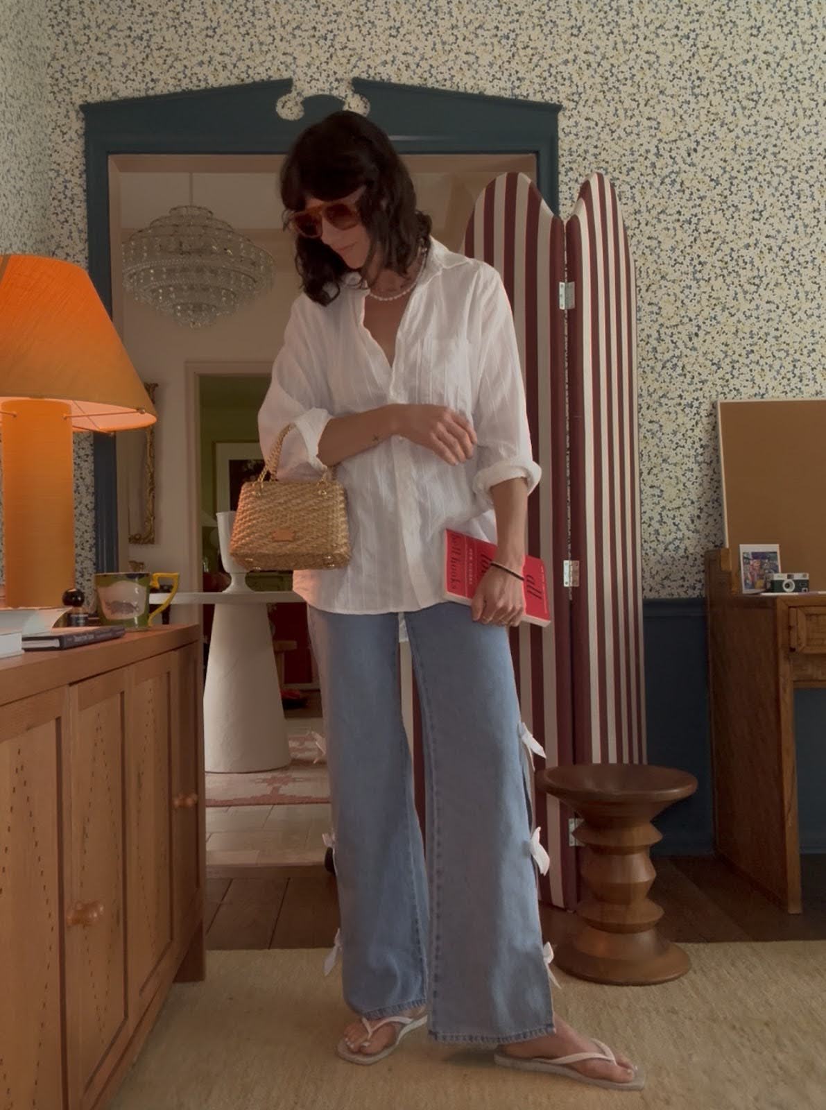 A woman wears flare jeans with bows on the sides, a white button-down shirt, flip flops, and a small woven bag