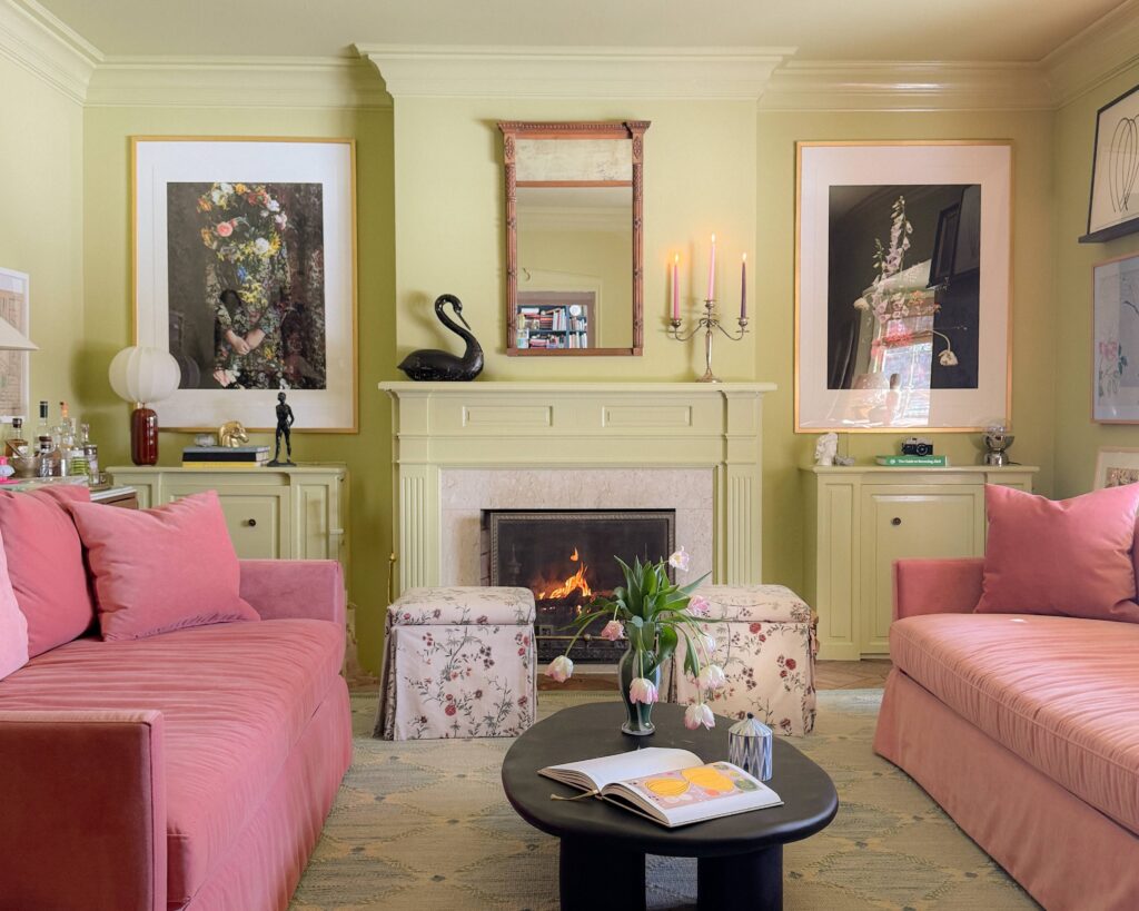 A green family room with pink velvet sofas, skirted ottomans, and a black oval coffee table