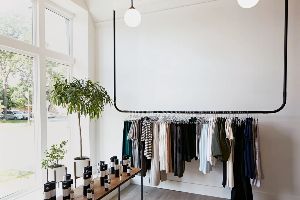 Idun St Paul with white walls, a clothing rack, perfumes, and plants in front of a large window