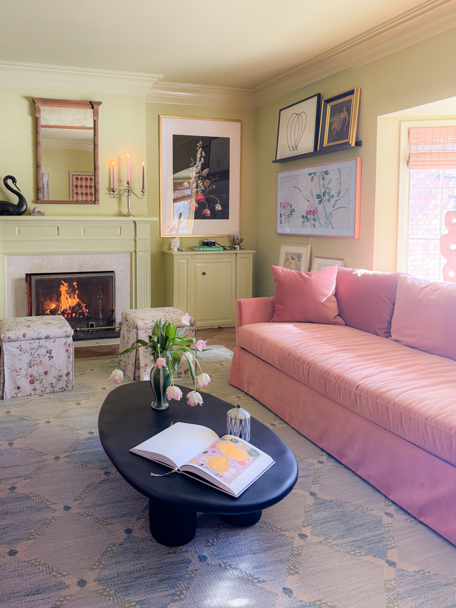 A green family room with pink velvet sofas, skirted ottomans, and a black oval coffee table