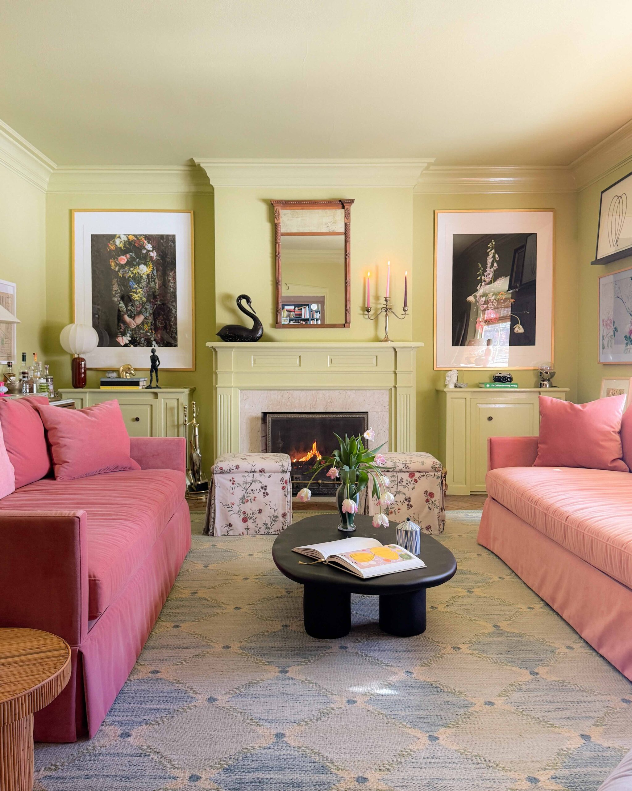 A green family room with pink velvet sofas, skirted ottomans, and a black oval coffee table