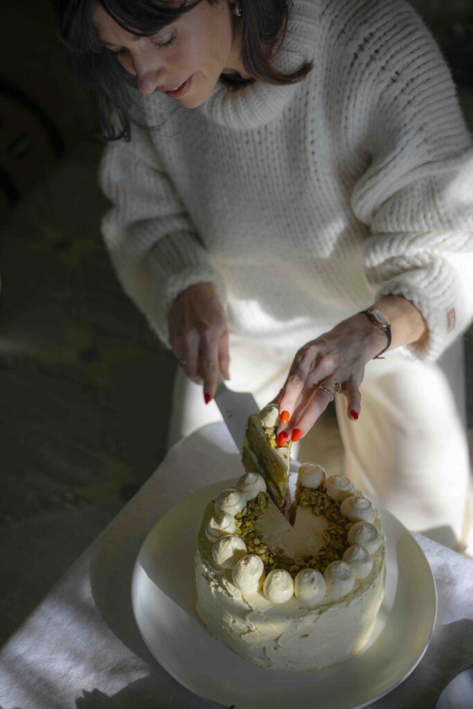 A woman wearing a cream-colored sweater and pants, cuts an olive oil ricotta cake