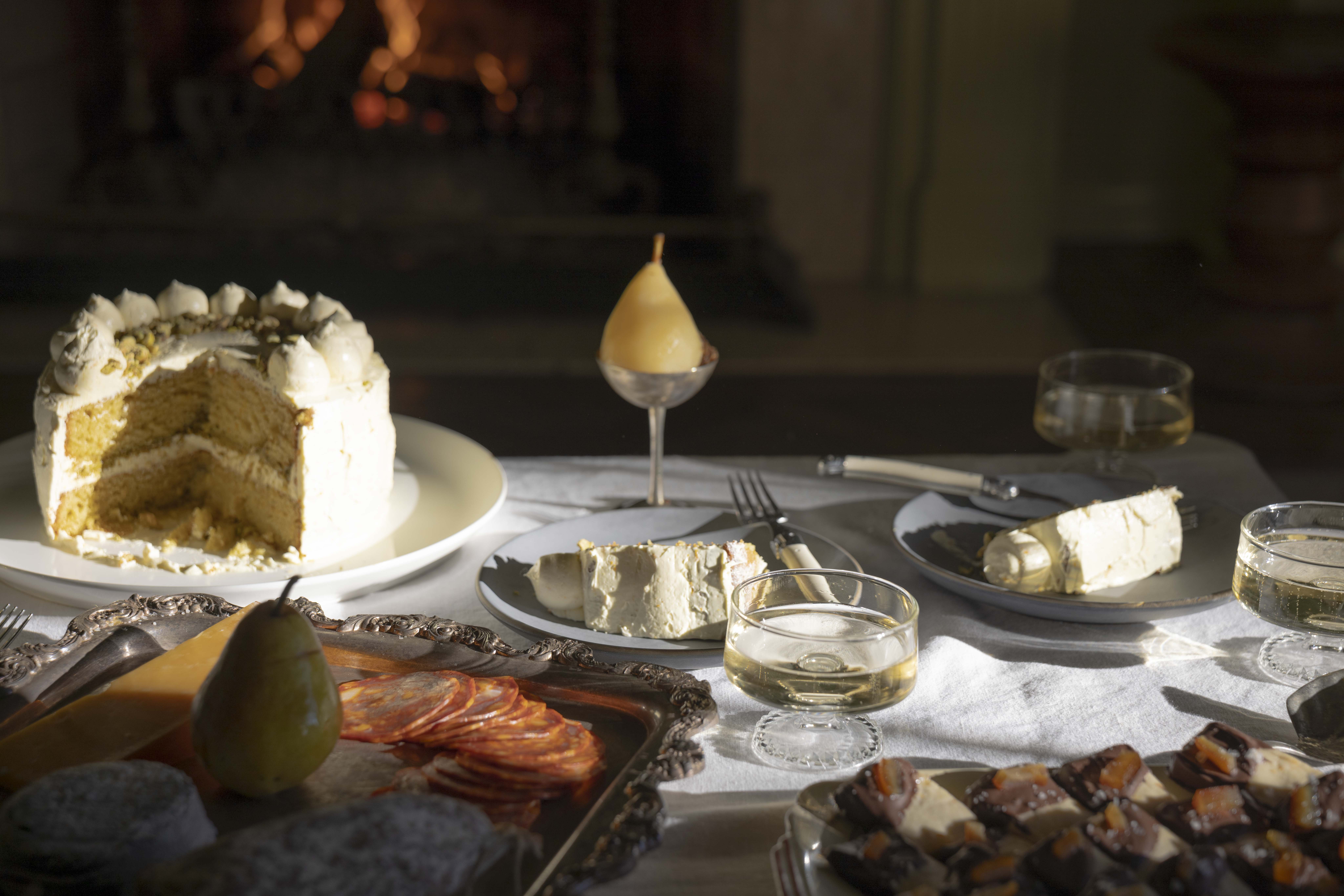 An olive oil ricotta cake sits on a plate on a coffee table, surrounded by charcuterie, shortbread, and glasses of sparkling wine